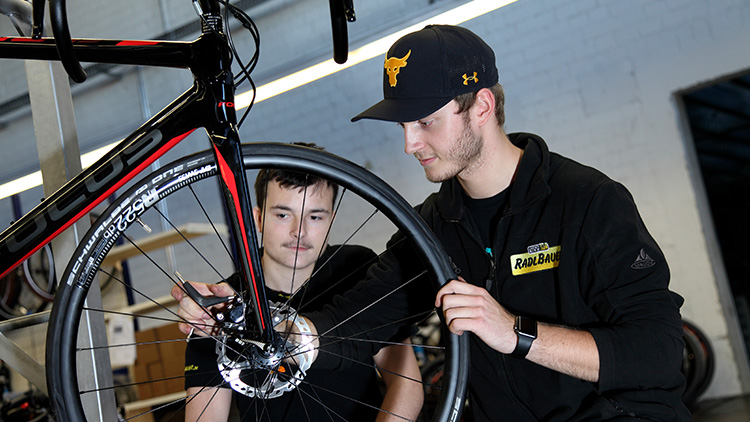 Zwei Junge Männer stehen hinter einem Fahrradreifen und reparieren es